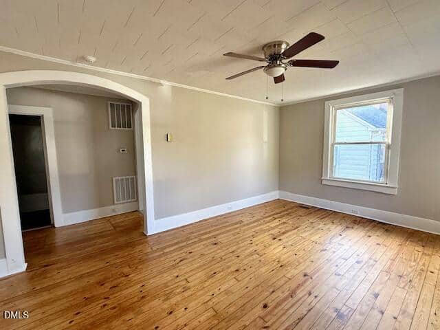Empty room with light-colored walls, a wooden floor, a ceiling fan with lights, a single window, and an arched doorway leading to another area. The room is well-lit by natural light.