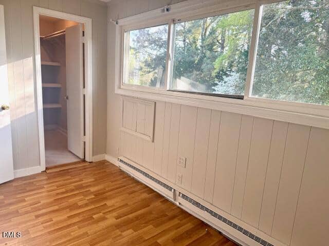 Small, empty room with light wood floors, paneled white walls, a large window with a view of trees, and an open door leading to a walk-in closet with shelves.