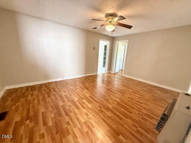 A spacious, empty room with light wood flooring, beige walls, a ceiling fan with lights, and two doorways leading to other rooms. The room is well-lit with natural and artificial light.
