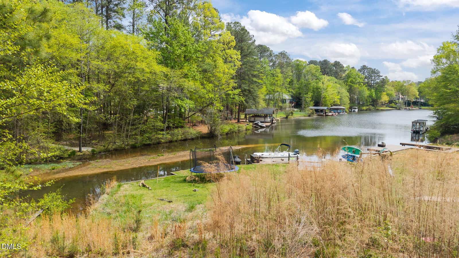 A small river or lake with boats docked along the shore, surrounded by green trees and grass. A trampoline is on the grassy area near the water, and several docks and houses are visible in the background.