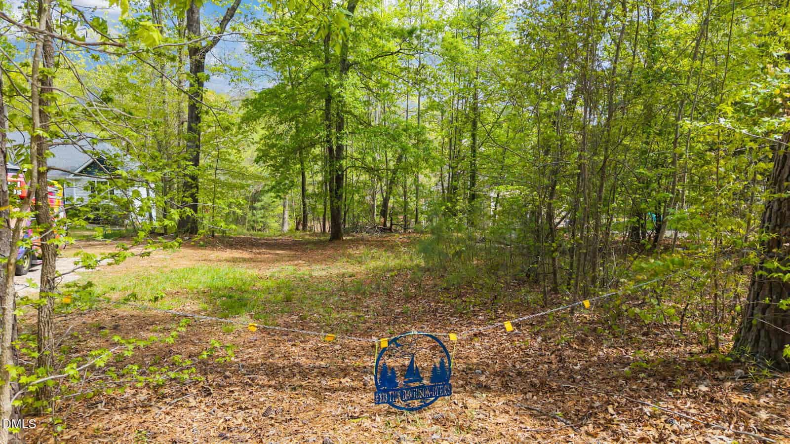 A wooded lot with green trees and leafy ground cover, bordered by a thin rope with a blue sign showing two trees. A house is partially visible on the left side.