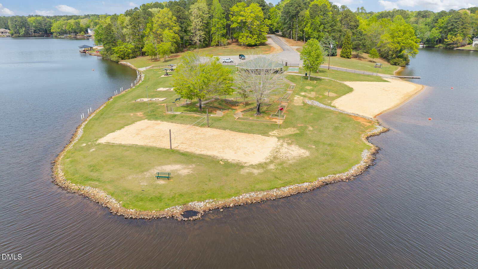 A small, sandy park on a lake features volleyball courts, benches, trees, and a parking lot. The area is surrounded by water, with forested land and houses visible in the background.