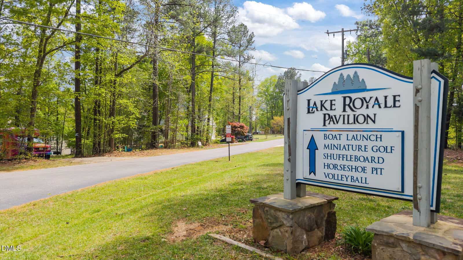 A sign for Lake Royale Pavilion lists amenities: boat launch, miniature golf, shuffleboard, horseshoe pit, and volleyball. It’s beside a road lined with trees on a sunny day.