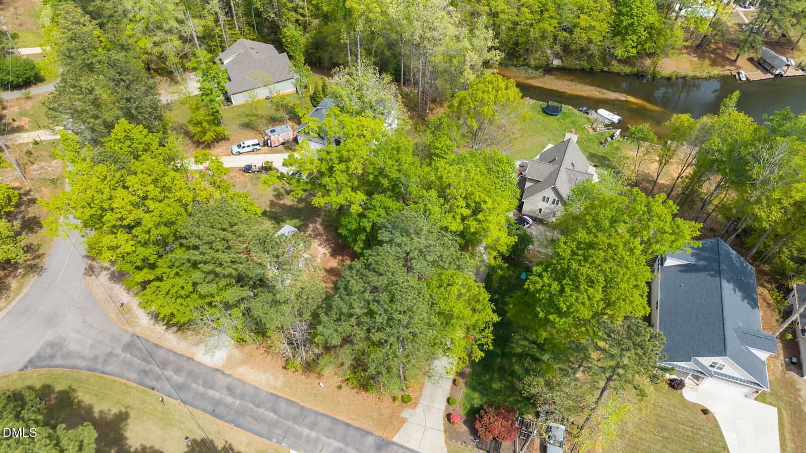 Aerial view of a suburban neighborhood with several houses surrounded by trees, a curved road, driveways, and a pond in the background. Some vehicles are parked near the homes. The area is lush and green with dense foliage.
