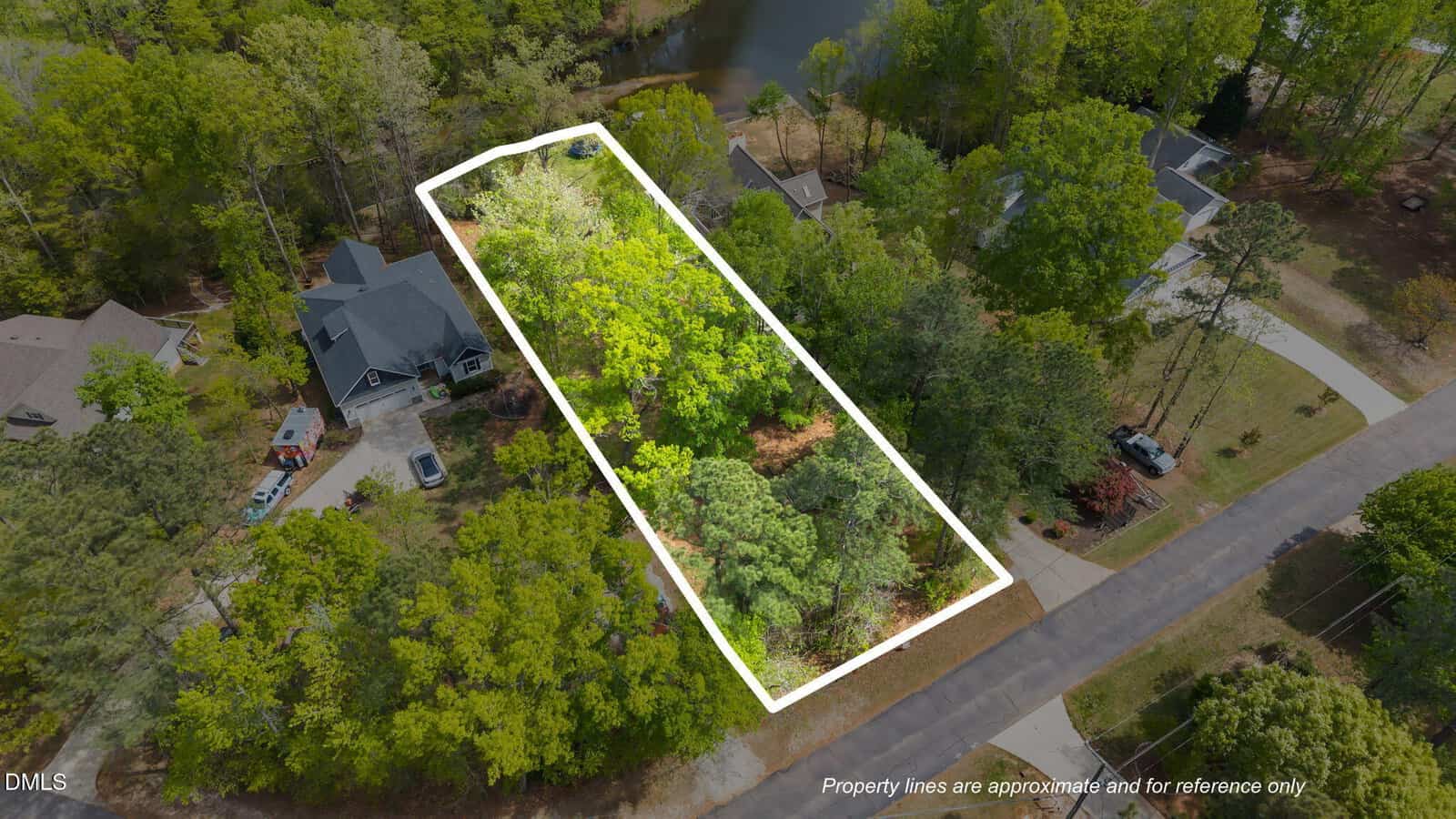 A rectangular, tree-covered vacant lot is outlined in white between two homes on a suburban street, with a pond visible behind the property. The area is surrounded by trees and other houses.