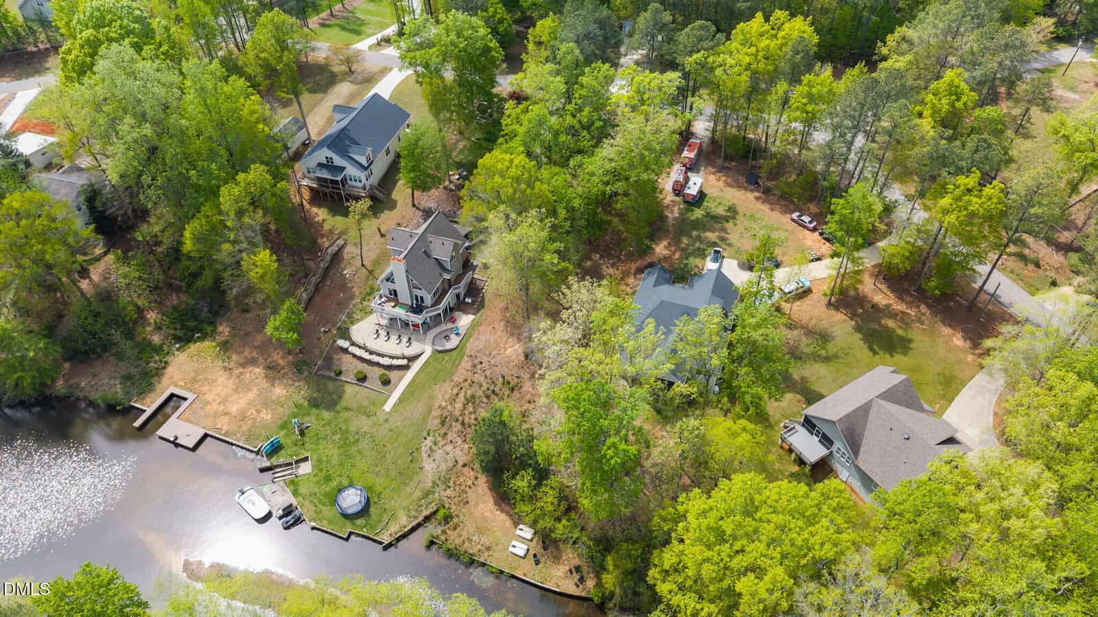 Aerial view of three houses surrounded by trees near a small lake with docks and boats in Louisburg. The homes, including one at 103 Chuckwagon Drive, are connected by driveways, with vehicles and outdoor furniture visible in the yards.