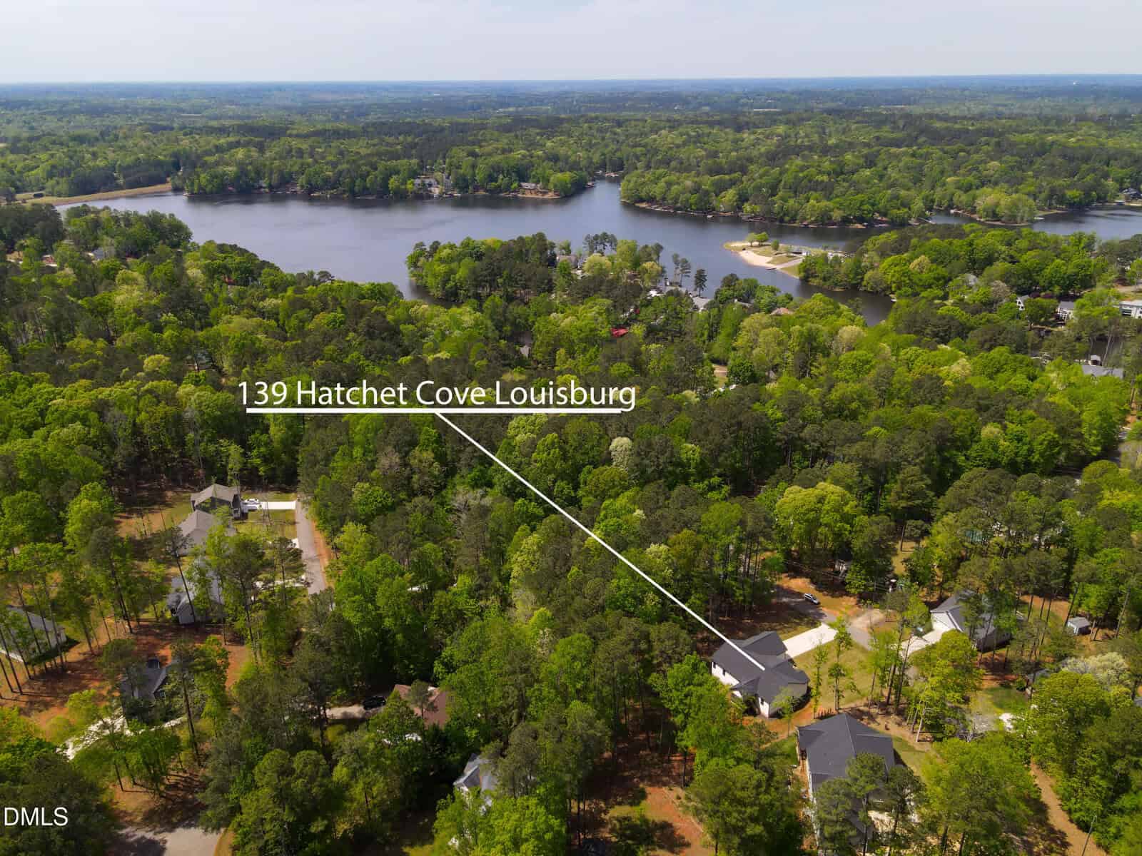 Aerial view of a wooded lakeside neighborhood with a house labeled “139 Hatchet Cove Louisburg” and a white line pointing to the property, trees and water in the background.