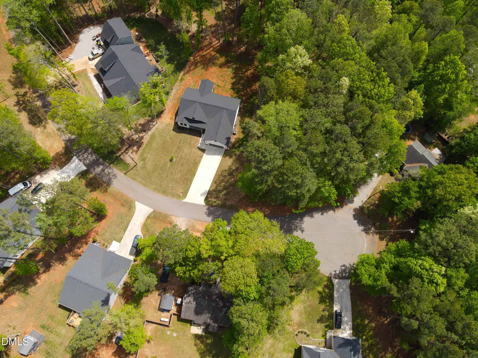Aerial view of a suburban neighborhood with several houses surrounded by trees. Curved roads and driveways connect the homes, which have lawns and some outdoor structures. The area is lush with greenery and appears well-maintained.