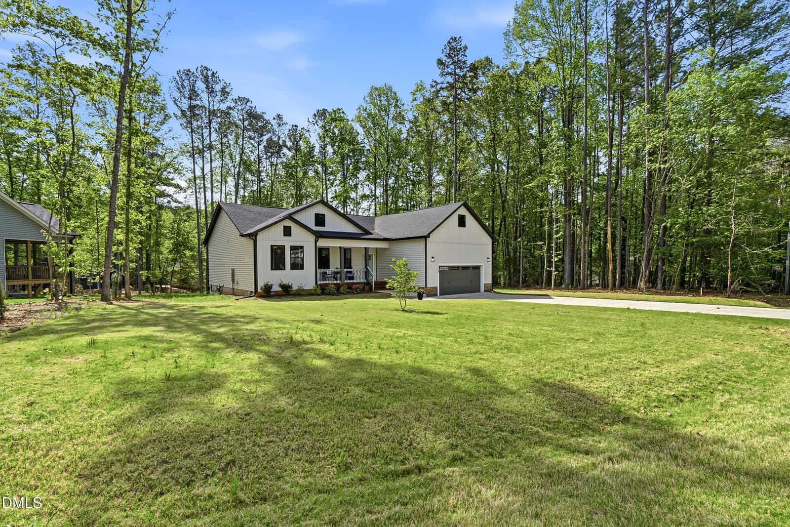 A single-story house with a front porch, attached two-car garage, and large grassy yard is surrounded by tall trees under a blue sky. A concrete driveway leads to the garage.