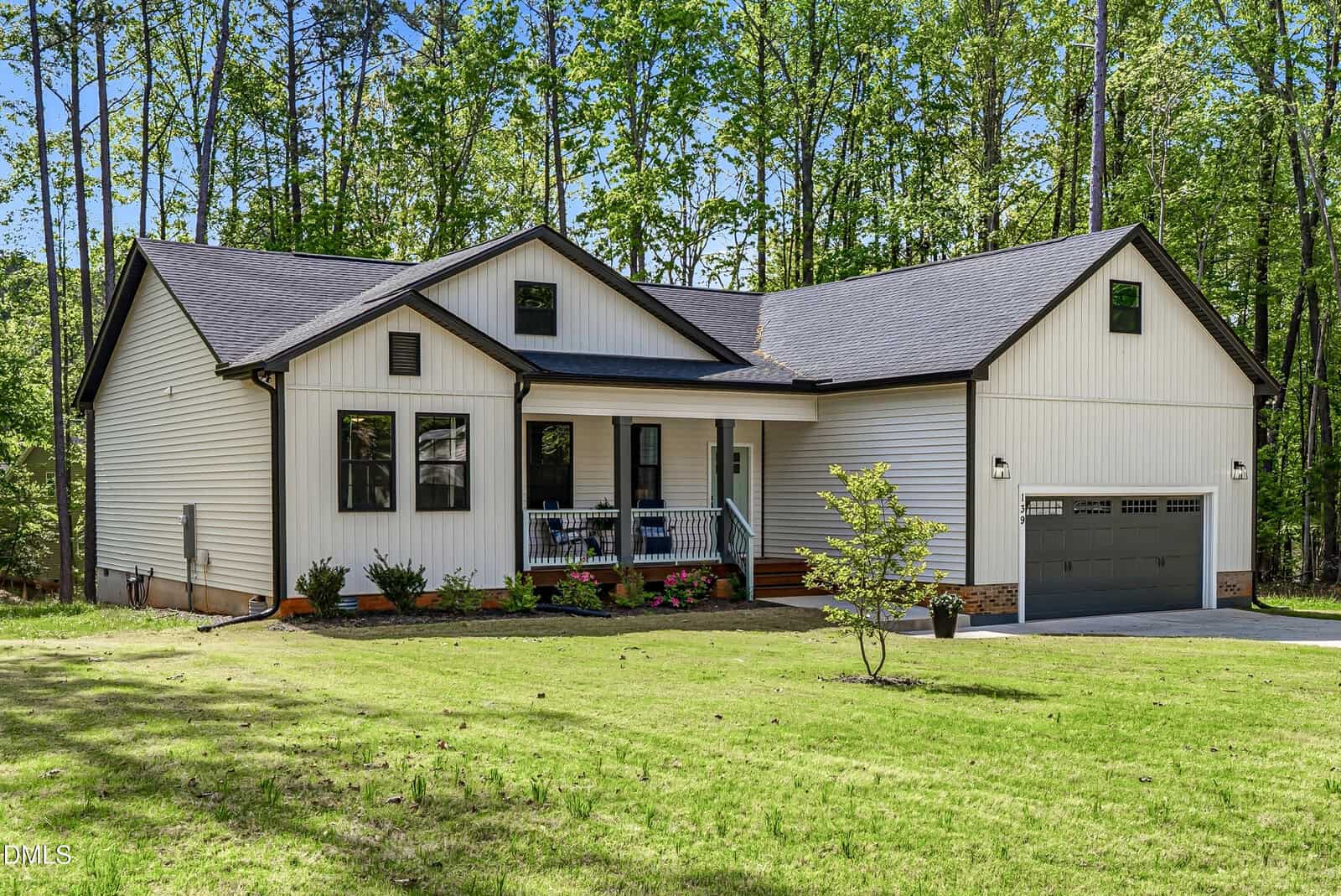 Single-story house with light-colored siding, front porch, dark roof, and two-car garage, set amid a green lawn and surrounded by tall trees. Small shrubs line the front of the house.