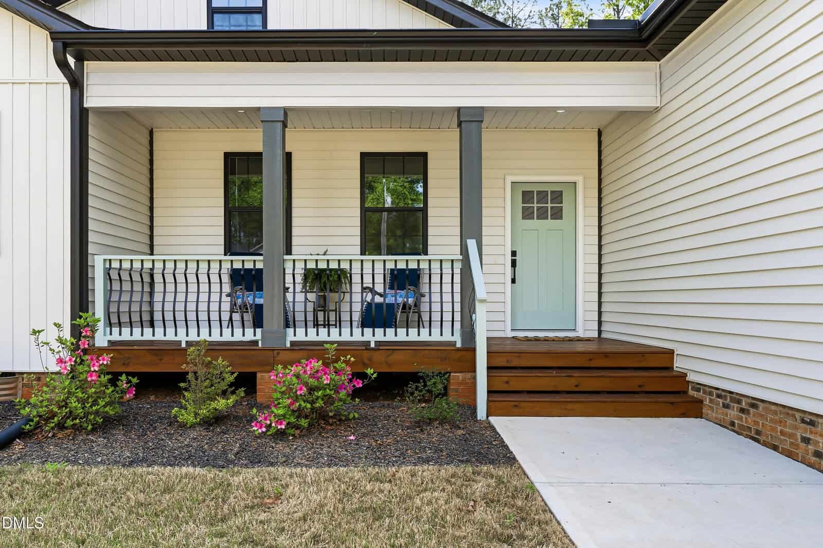 A modern house entrance with a mint green door, two large windows, a small porch with dark railings, two chairs, potted plants, wooden steps, and blooming bushes in a landscaped front yard.