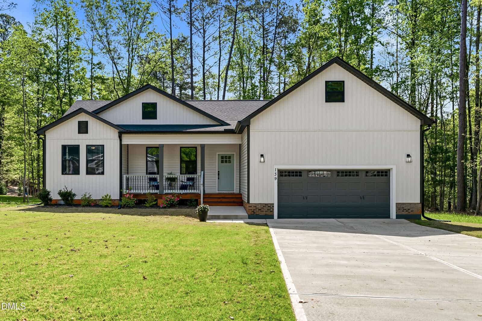 Single-story house with white vertical siding, black trim, a covered front porch with steps, landscaped flower beds, a two-car garage, and a concrete driveway, surrounded by green grass and tall trees in the background.