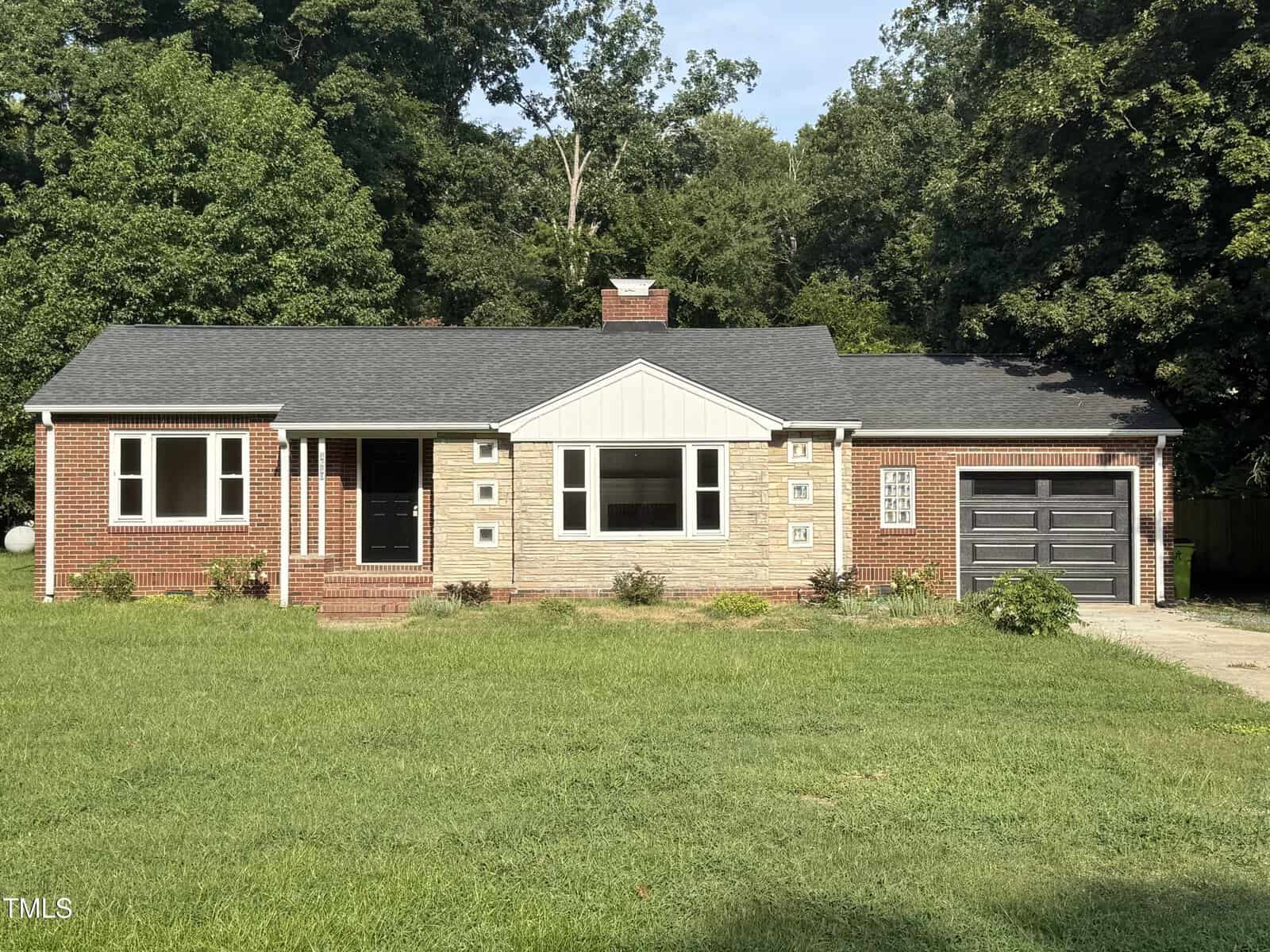 Charming single-story brick house with a black roof, attached garage, and white-framed windows at 1610 W B Street in Butner. A central front door welcomes you home as the house sits on a grassy lawn, bordered by trees in the background.