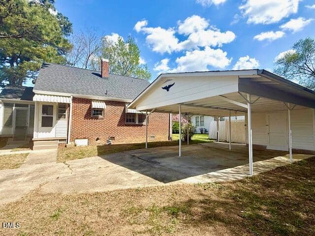 Single-story brick house with a dark gray roof, covered carport featuring a bird decoration, concrete driveway, and a small backyard area with trees and a partly cloudy sky.