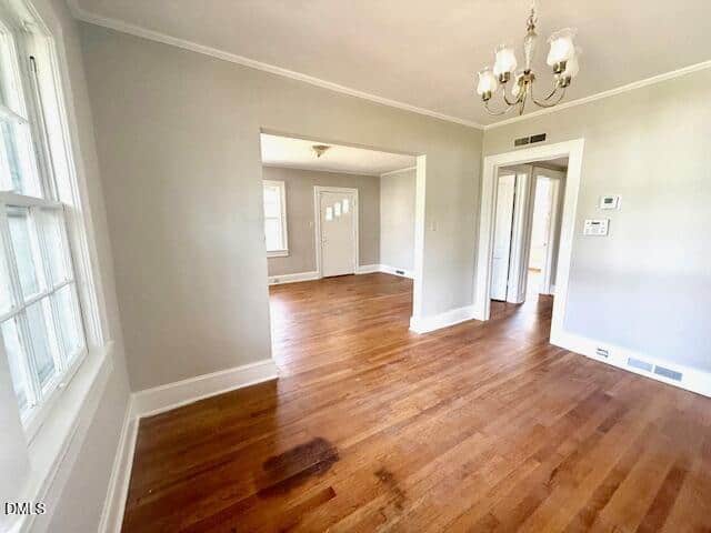 Bright, empty room with hardwood floors, light gray walls, white trim, a window on the left, and a chandelier. Doorways lead to other rooms, one with a white exterior door and another with a ceiling light.