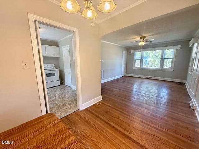 A bright room with wood floors, an overhead light, an adjacent living area with a ceiling fan, and a view into a white kitchen with tile flooring. Large windows let in natural light.