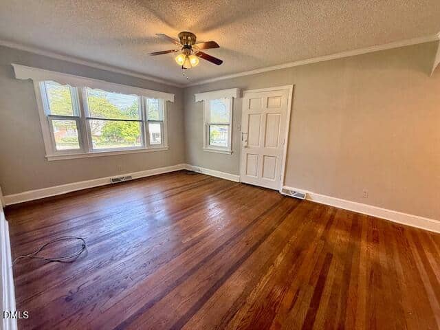 Bright, empty room with hardwood floors, beige walls, a ceiling fan with lights, three large windows letting in natural light, and a white closet door. A black cable lies on the floor near the windows.