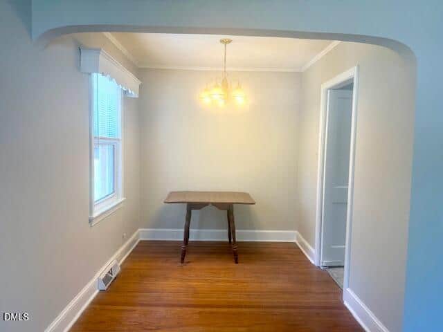 Small dining nook with light gray walls, a single window with a valance, wooden floors, a vintage wooden table, and a ceiling light fixture with three bulbs. An open doorway is on the right side.