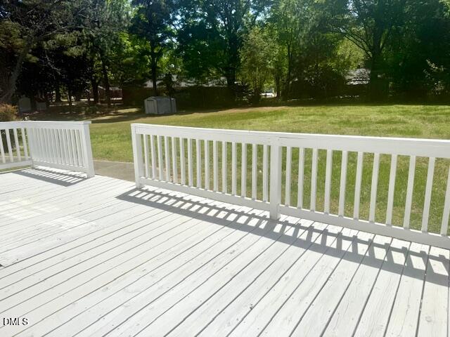 A sunlit wooden deck with white railings overlooks a large, grassy backyard with trees and a storage shed in the distance. Shadows from the railings fall across the deck.