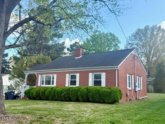 A single-story red brick house with white trim, a black roof, and multiple windows. Neatly trimmed bushes line the front, and there is a grassy lawn and trees in the background under a blue sky.