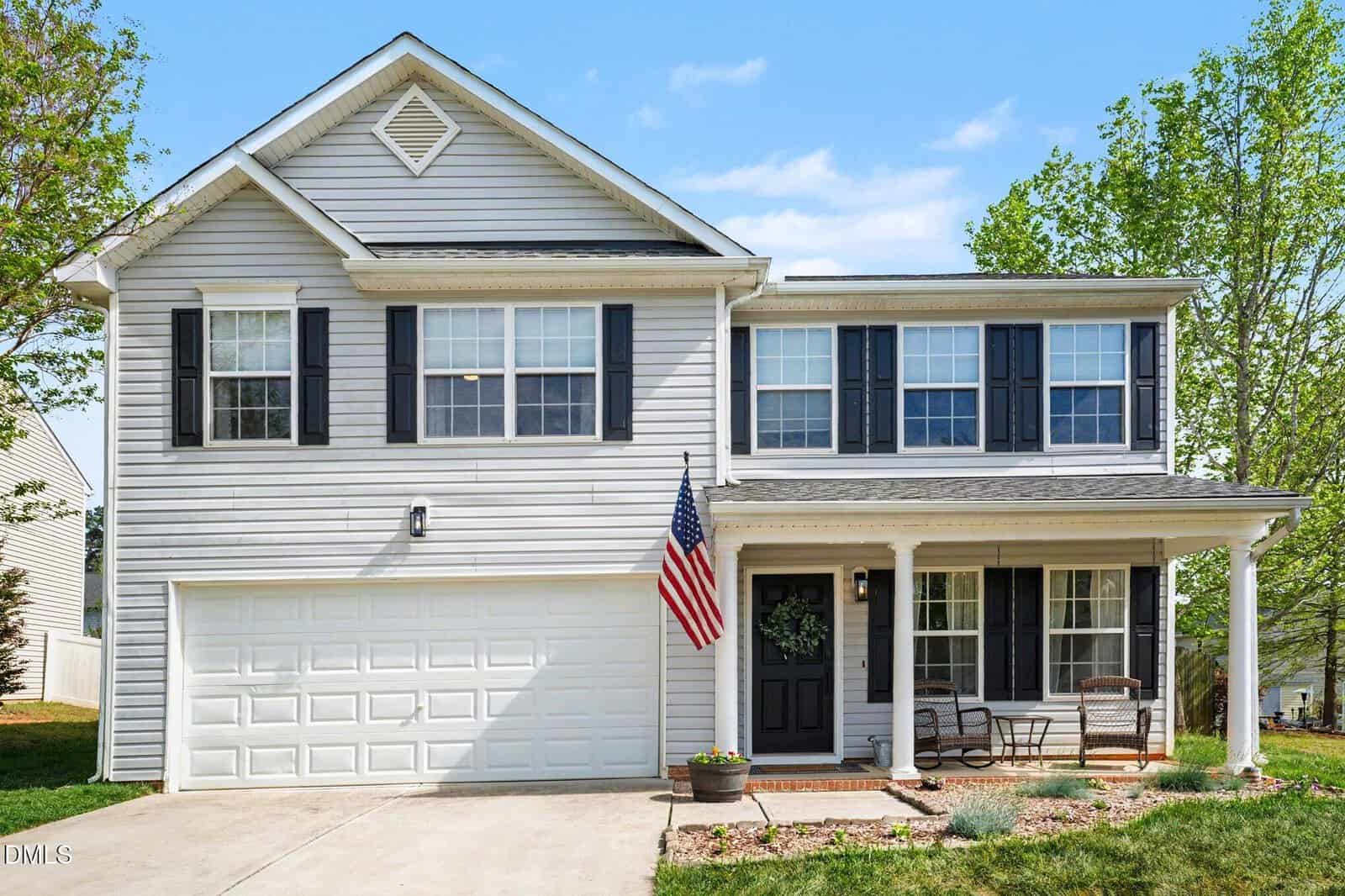 Two-story white house with black shutters, double garage, front porch with chairs, an American flag, and a wreath on the door. Located at 704 Stackhurst Way in Wake Forest, this charming home is surrounded by a green lawn and trees under a clear blue sky.