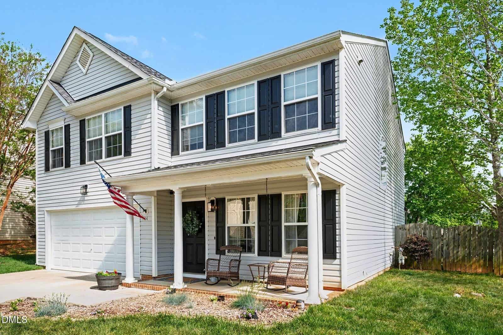 Two-story gray house with black shutters, front porch with two chairs, American flag, attached garage, and a small landscaped yard with a wooden fence and green trees under a clear blue sky.
