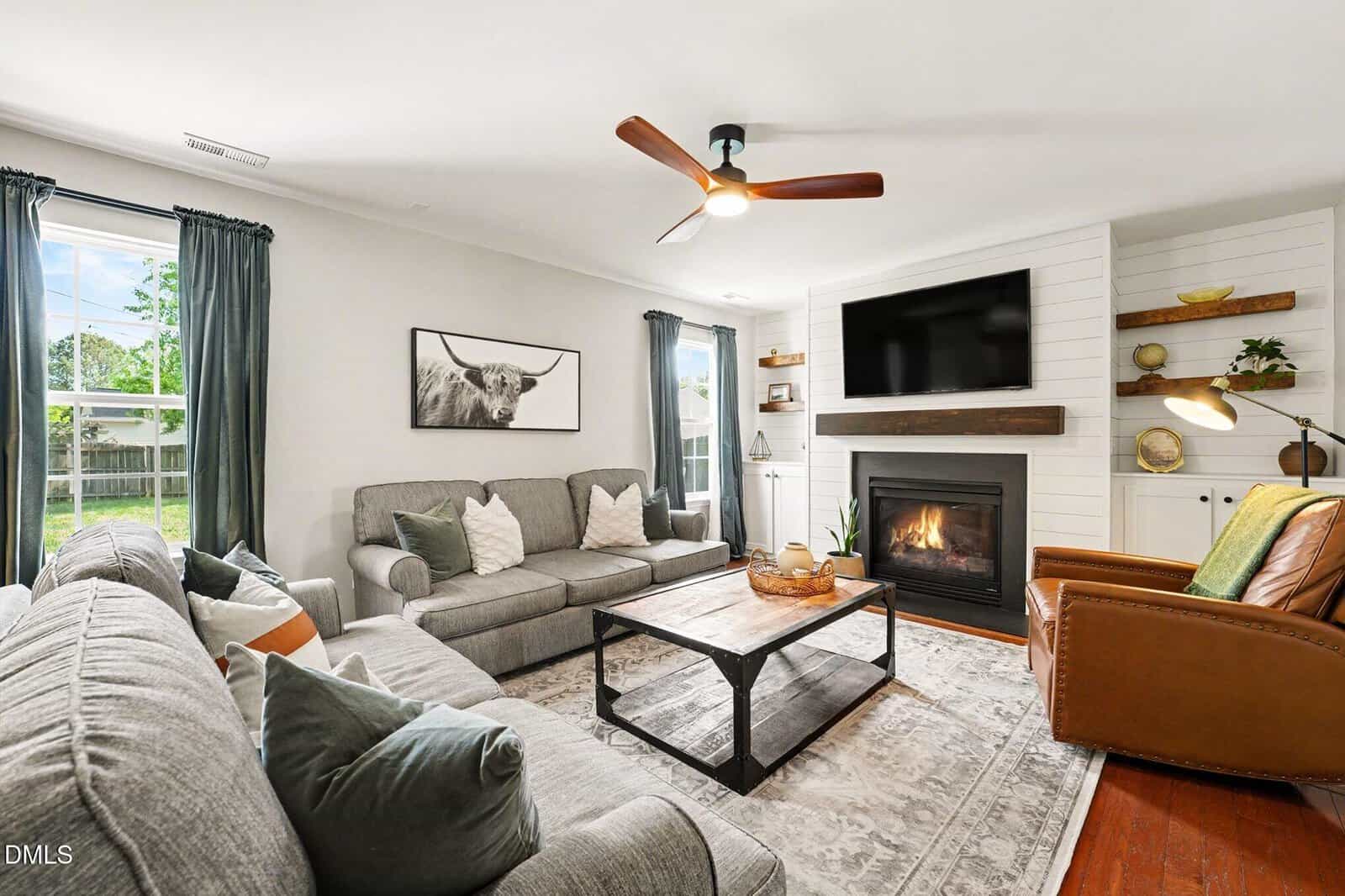 Bright living room with gray sofas, brown leather chair, fireplace, mounted TV, large window, blue curtains, and a coffee table on a patterned rug. Decor includes shelves, art, and plants.