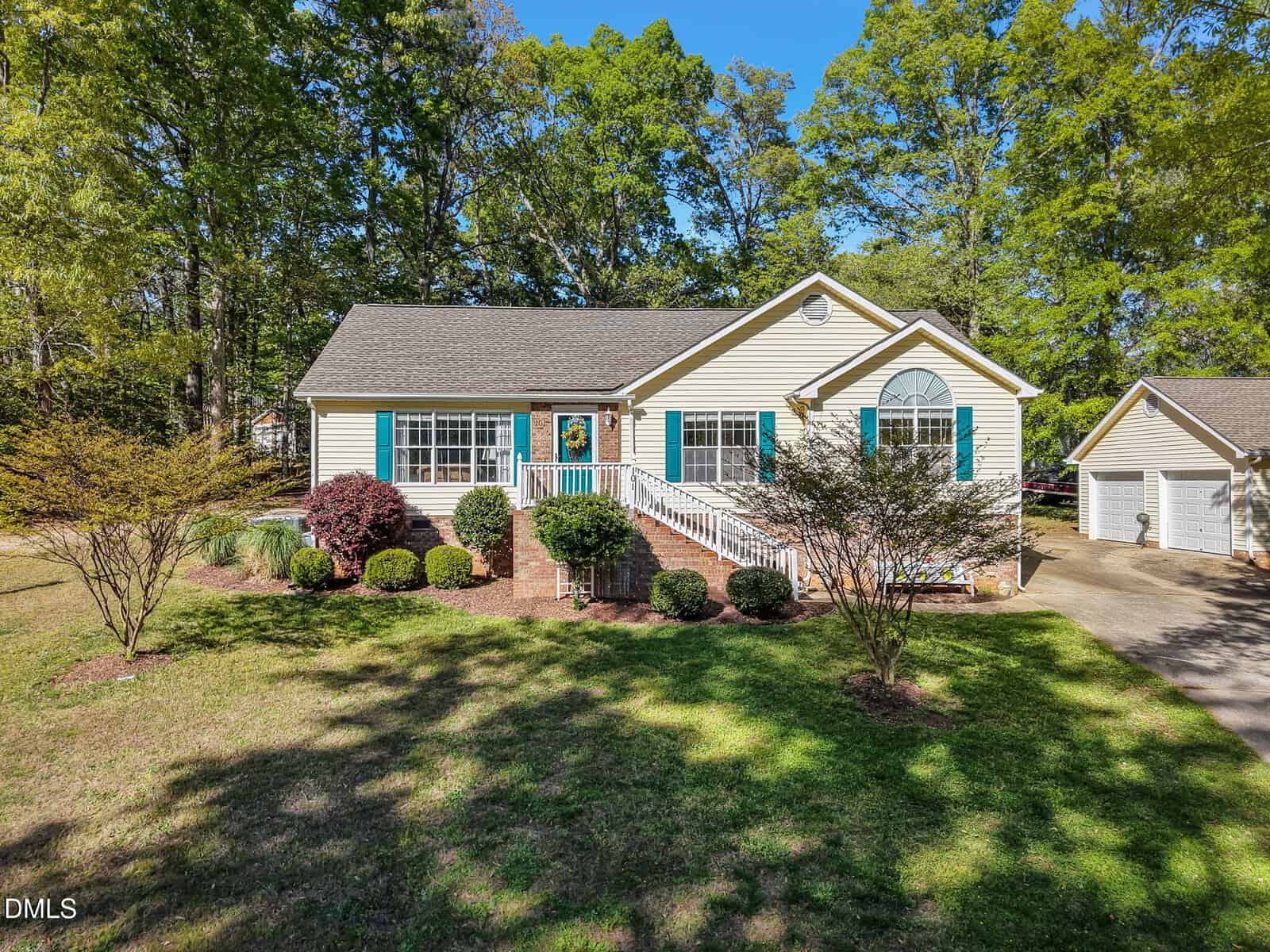 Charming one-story beige house in Louisburg with blue shutters, a welcoming front porch framed by white railings, landscaped shrubs, and a driveway leading to a detached garage at 101 Stage Line Cove—nestled among green lawns and trees under a clear blue sky.