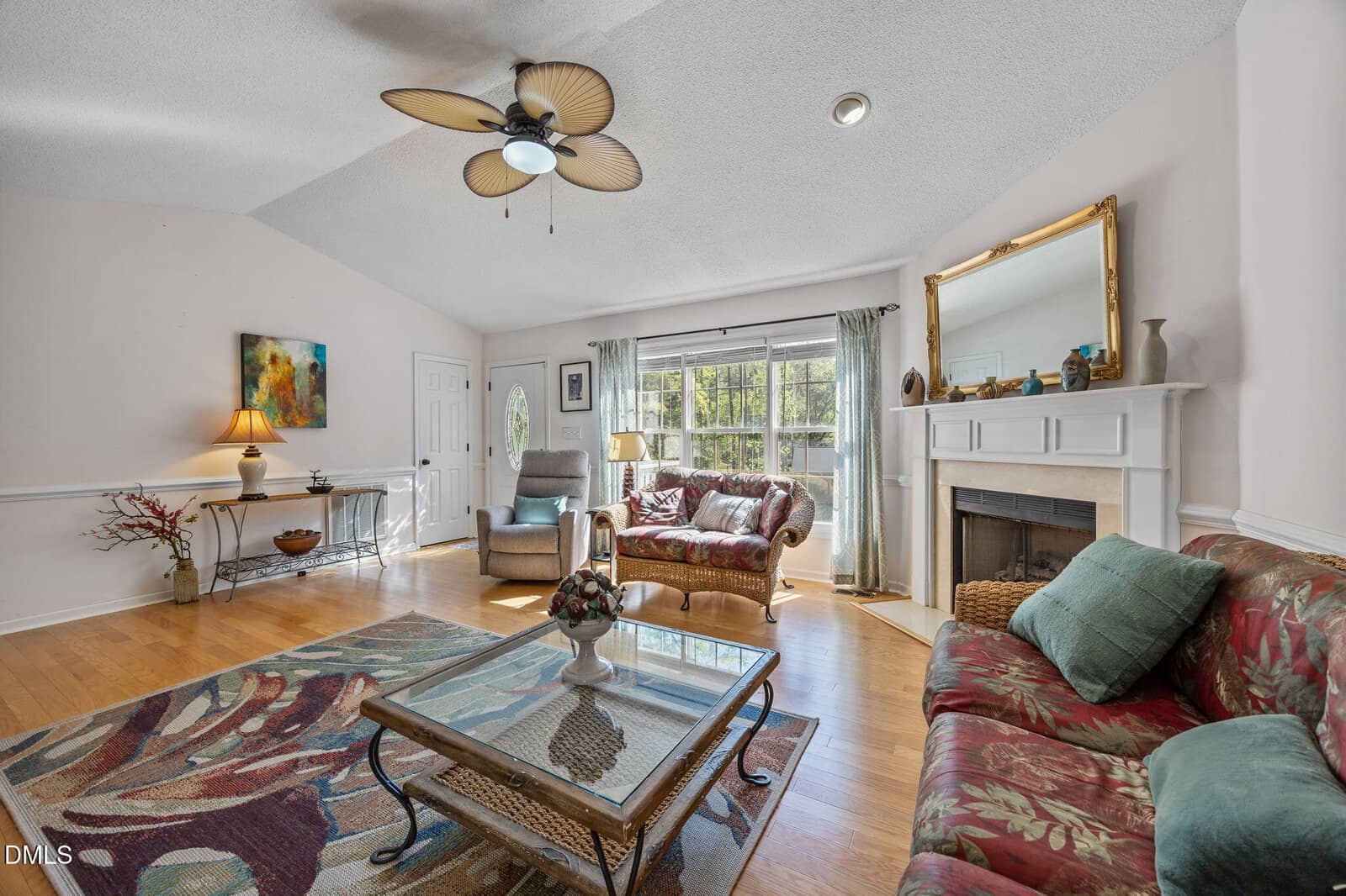Bright living room with hardwood floors, two patterned sofas, glass coffee table, fireplace with mirror, large window with sheer curtains, ceiling fan, and decorative accents, all under a vaulted ceiling.