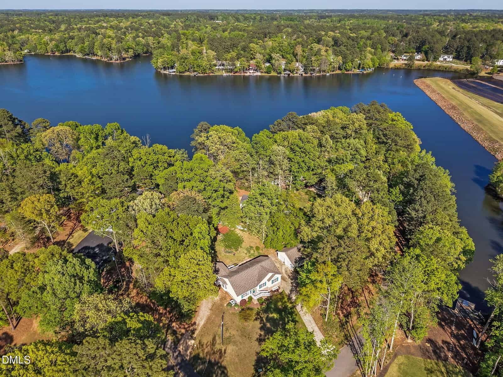 Aerial view of a house surrounded by trees near a large lake, with lush green forest and shoreline visible in the background.