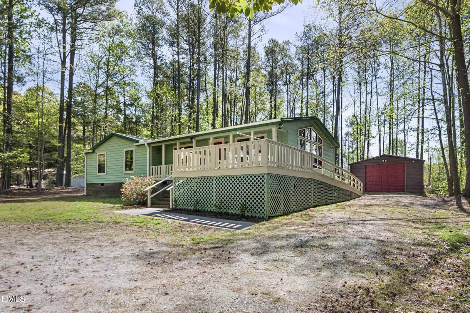Single-story green house with a front porch, ramp, and lattice skirting sits among tall trees. A red detached garage is visible in the background, and the driveway is gravel with some grass and dirt patches.
