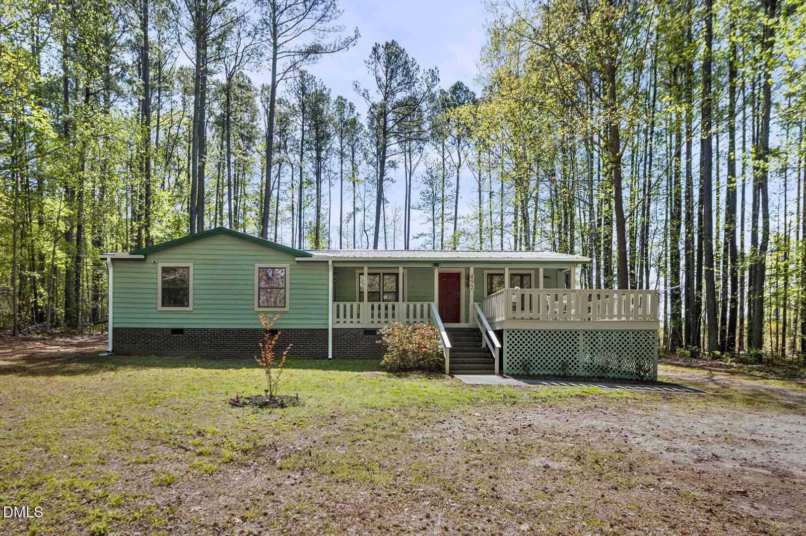 Charming single-story home at 492 Little Mill Road, Henderson, with light green siding, green trim, and a striking red front door. Features a covered front porch with white railings, brick foundation, and stairs. Surrounded by tall trees with a grassy front yard.