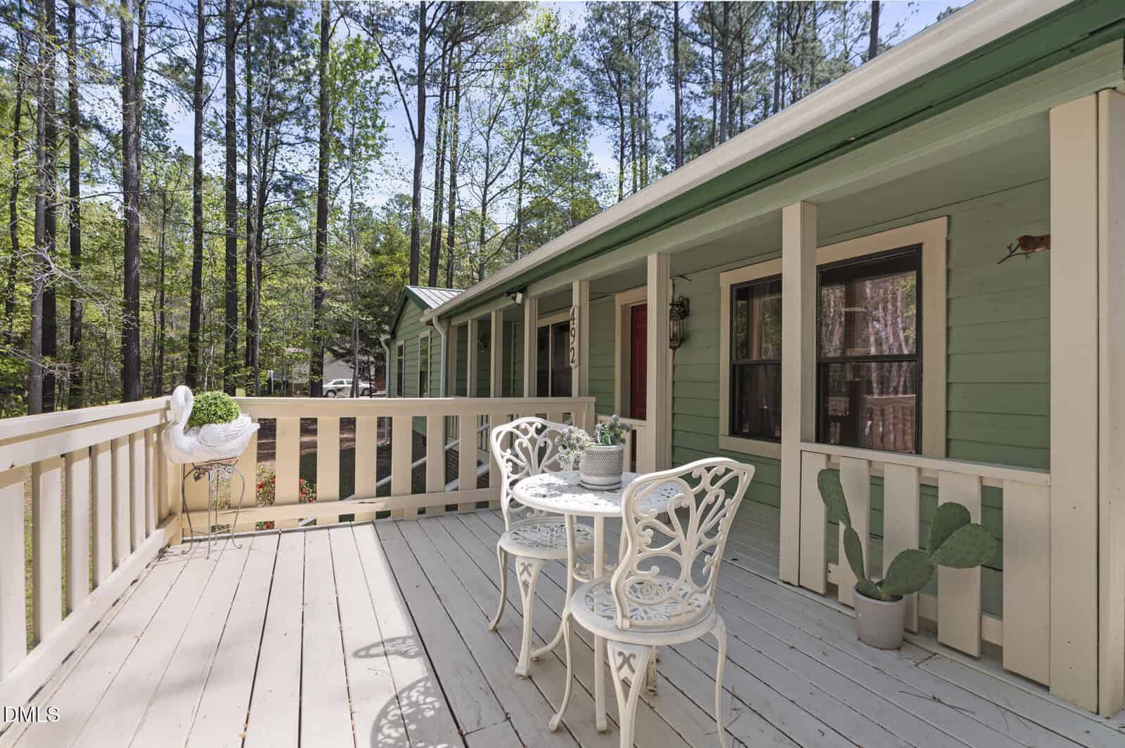 A pale green house with a covered porch, white railing, and two white metal chairs with a matching round table. A swan planter and a cactus in a pot sit near the railing, surrounded by tall pine trees.