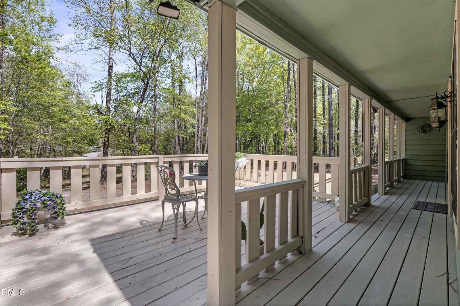 A wooden porch with a railing, a small table and two chairs, potted plants, and a forested area in the background. The porch is covered and receives natural light from the side.