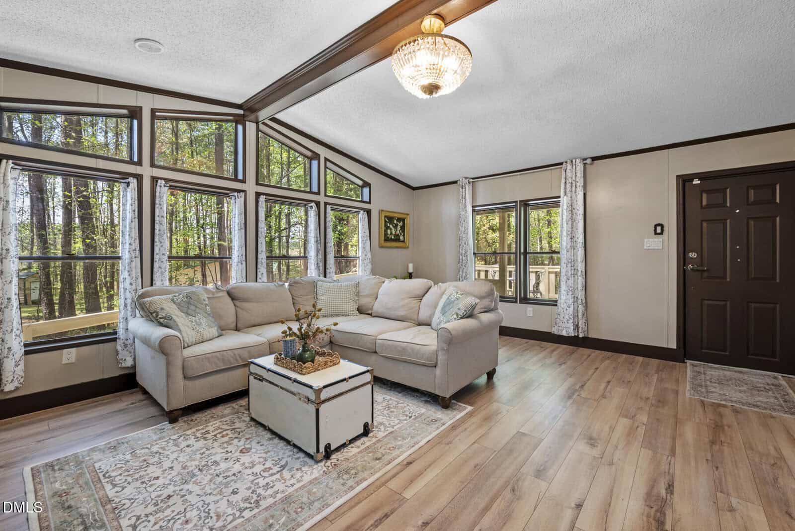 Bright living room with large corner windows, beige sectional sofa, patterned rug, vintage trunk coffee table, wood floors, and a chandelier. Neutral tones and forest view outside create a cozy and inviting atmosphere.