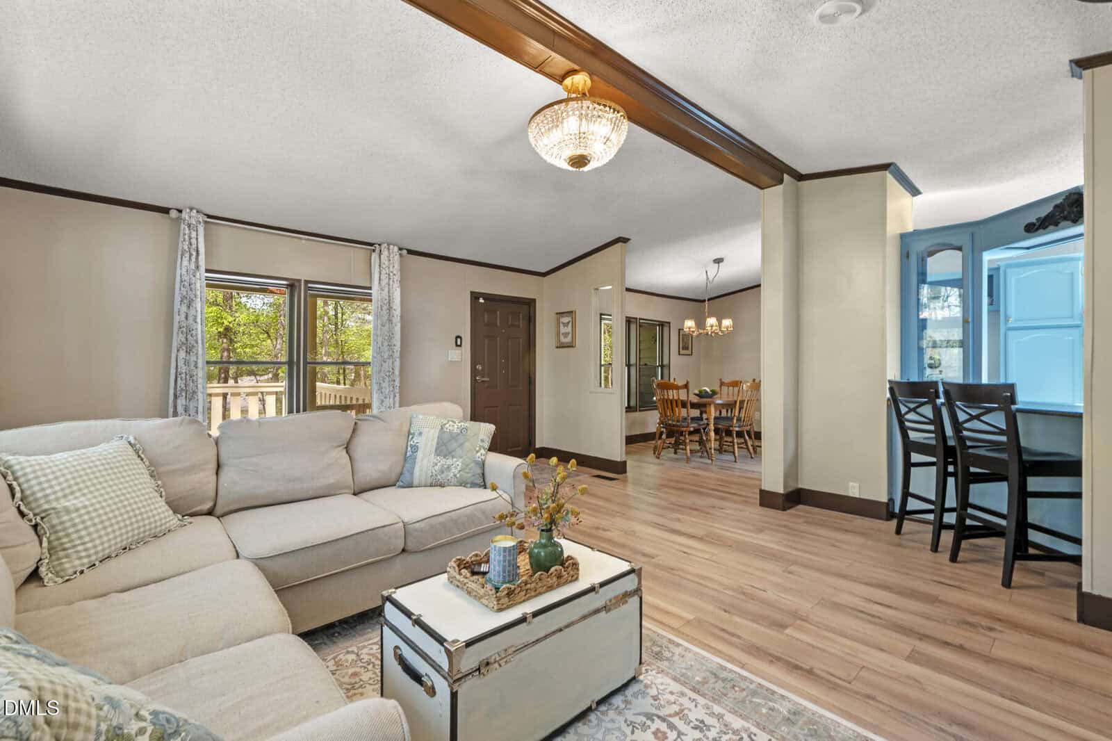 Bright living room with a beige sectional sofa, wooden floors, and a white chest as a coffee table, opening to a dining area and kitchen with light blue cabinets and bar stools. Large windows let in natural light.