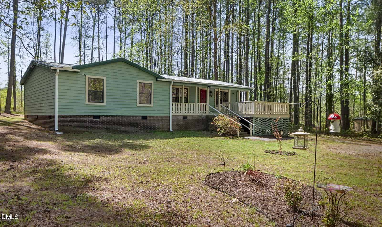 Single-story green house with white trim, brick foundation, and front porch. Surrounded by tall trees, the yard features simple landscaping, a bird feeder, and a mulched garden bed in the foreground.