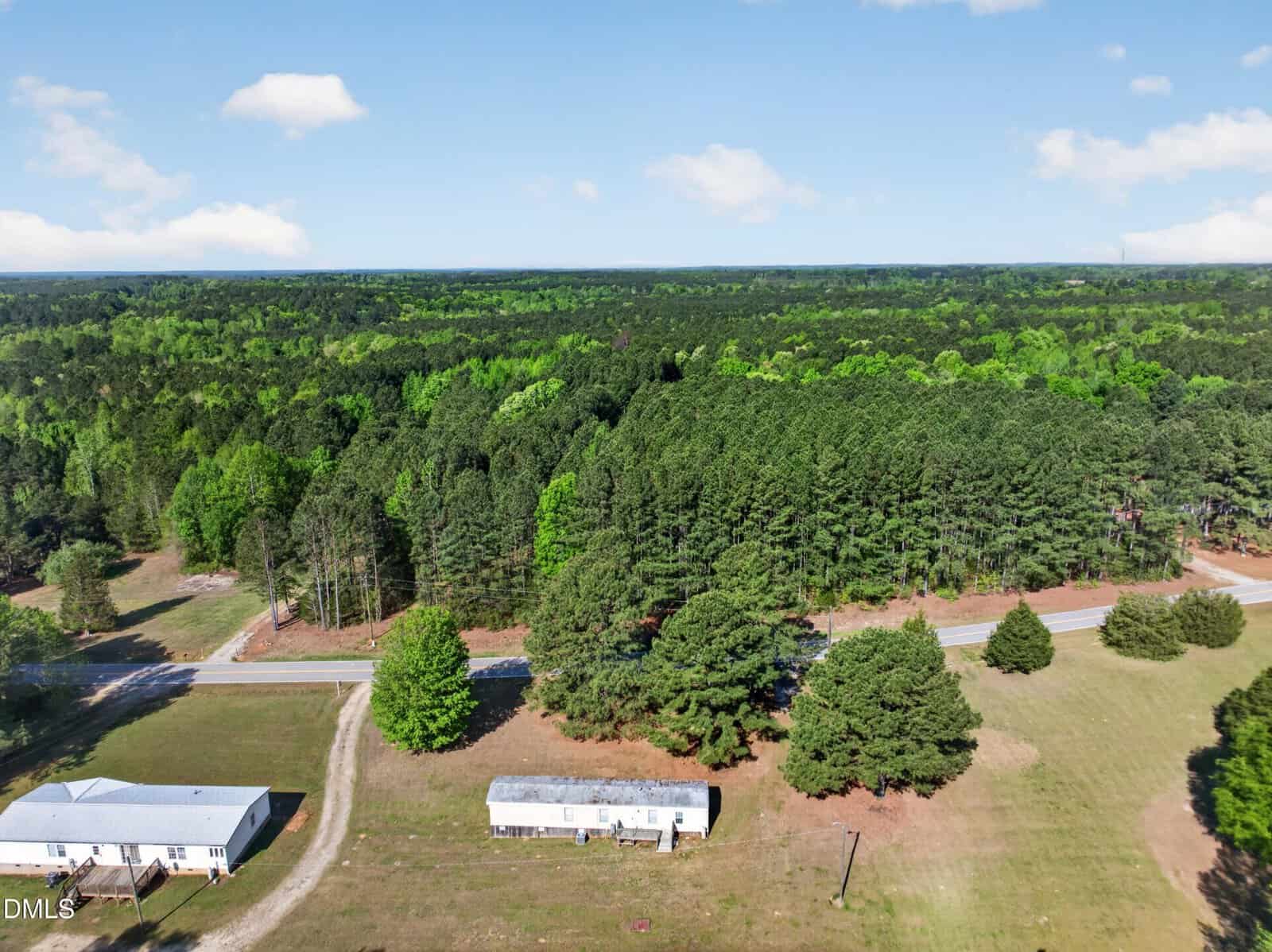 Aerial view of a rural area with a white house, a mobile home, open grassy land, and a road bordered by dense green forest under a blue sky with scattered clouds.