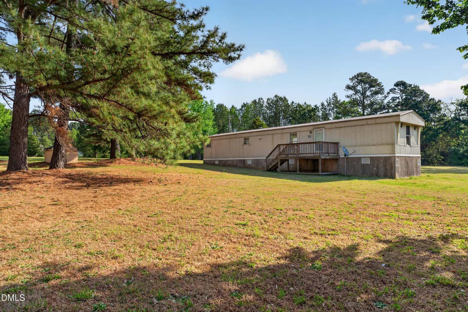 A beige mobile home with a small porch sits on a grassy, partially shaded lot with tall trees and clear blue sky in the background at 495 Macon-Embro Road, just outside Macon.