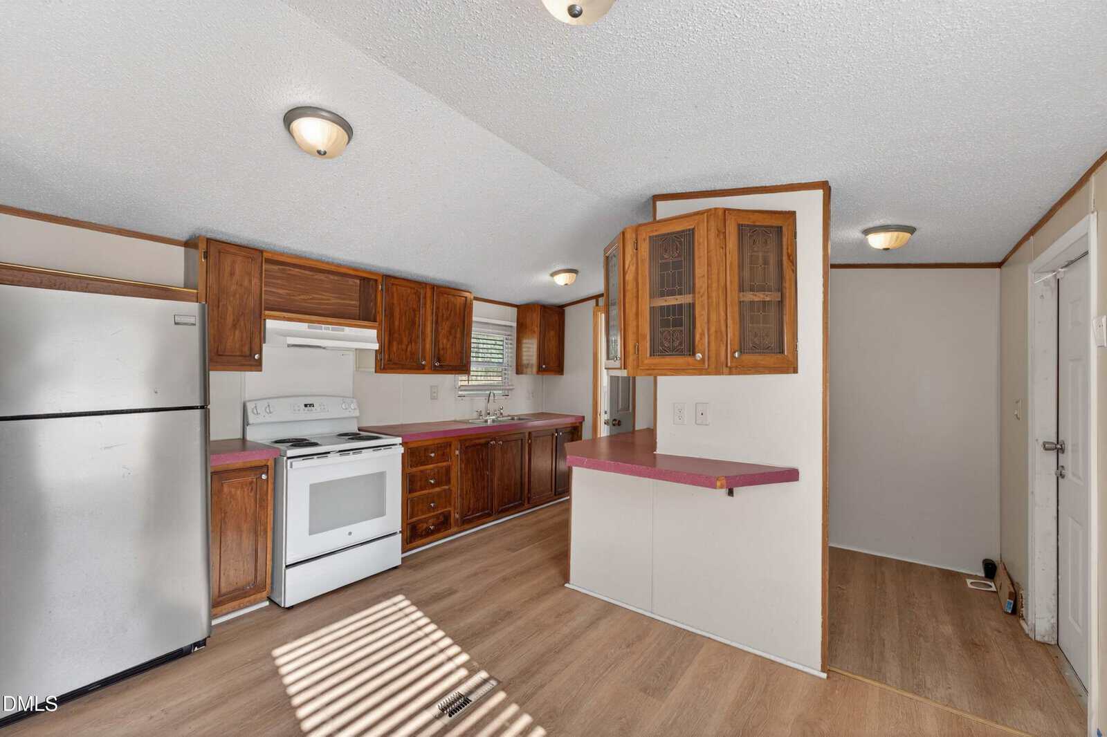 Kitchen with wooden cabinets, white appliances, pink countertops, and light wood flooring. There’s a stainless steel fridge, a window above the sink, and a small breakfast bar with display cabinets above.