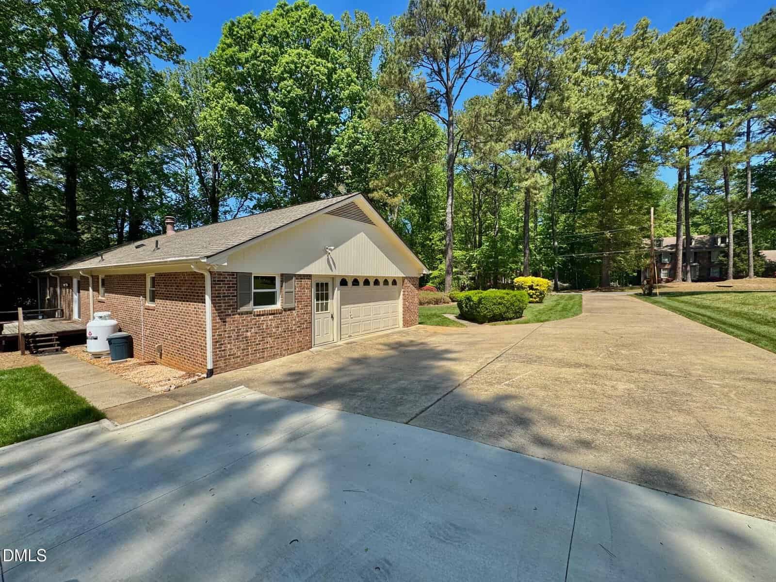 Single-story brick house with a three-car garage, wide concrete driveway, green lawn, bushes, and tall trees in the background under a clear blue sky.