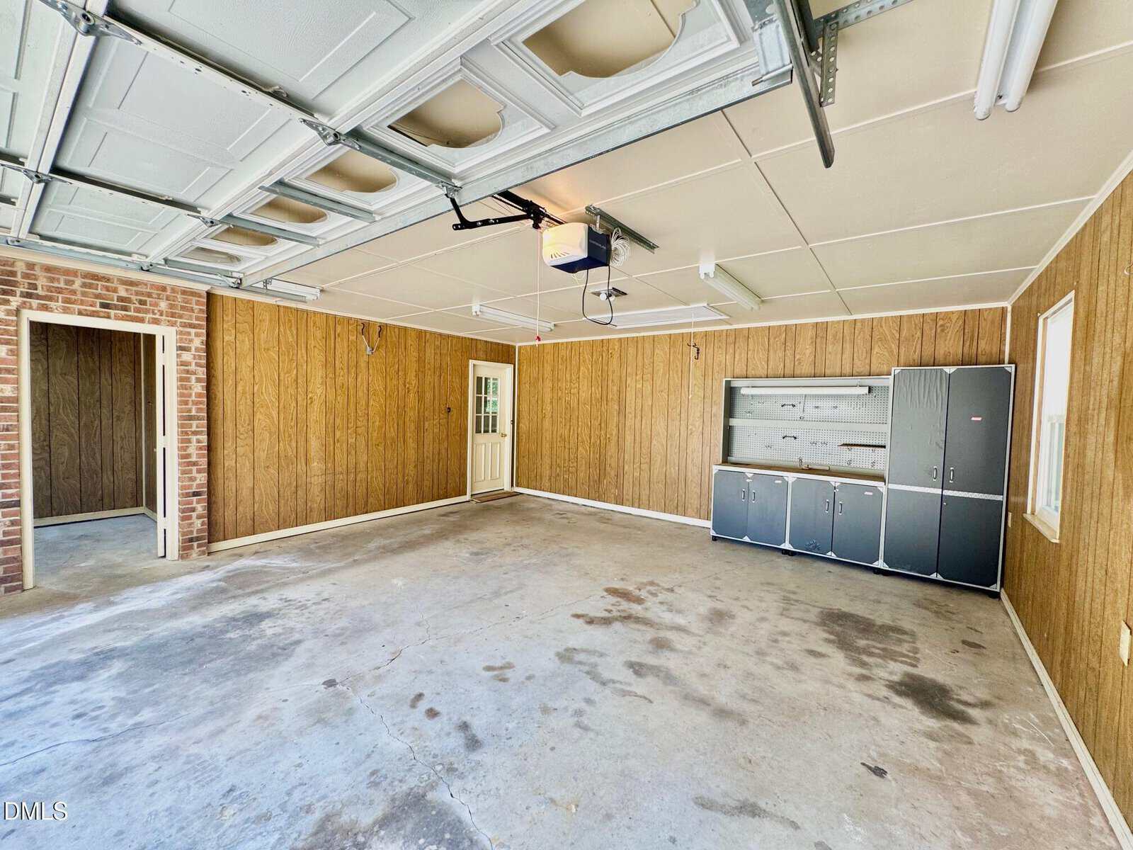 Spacious garage with wood-paneled walls, concrete floor, overhead garage door, storage cabinets, a door to the outside, and a doorway to an interior room. Brightly lit with ceiling lights and windows.
