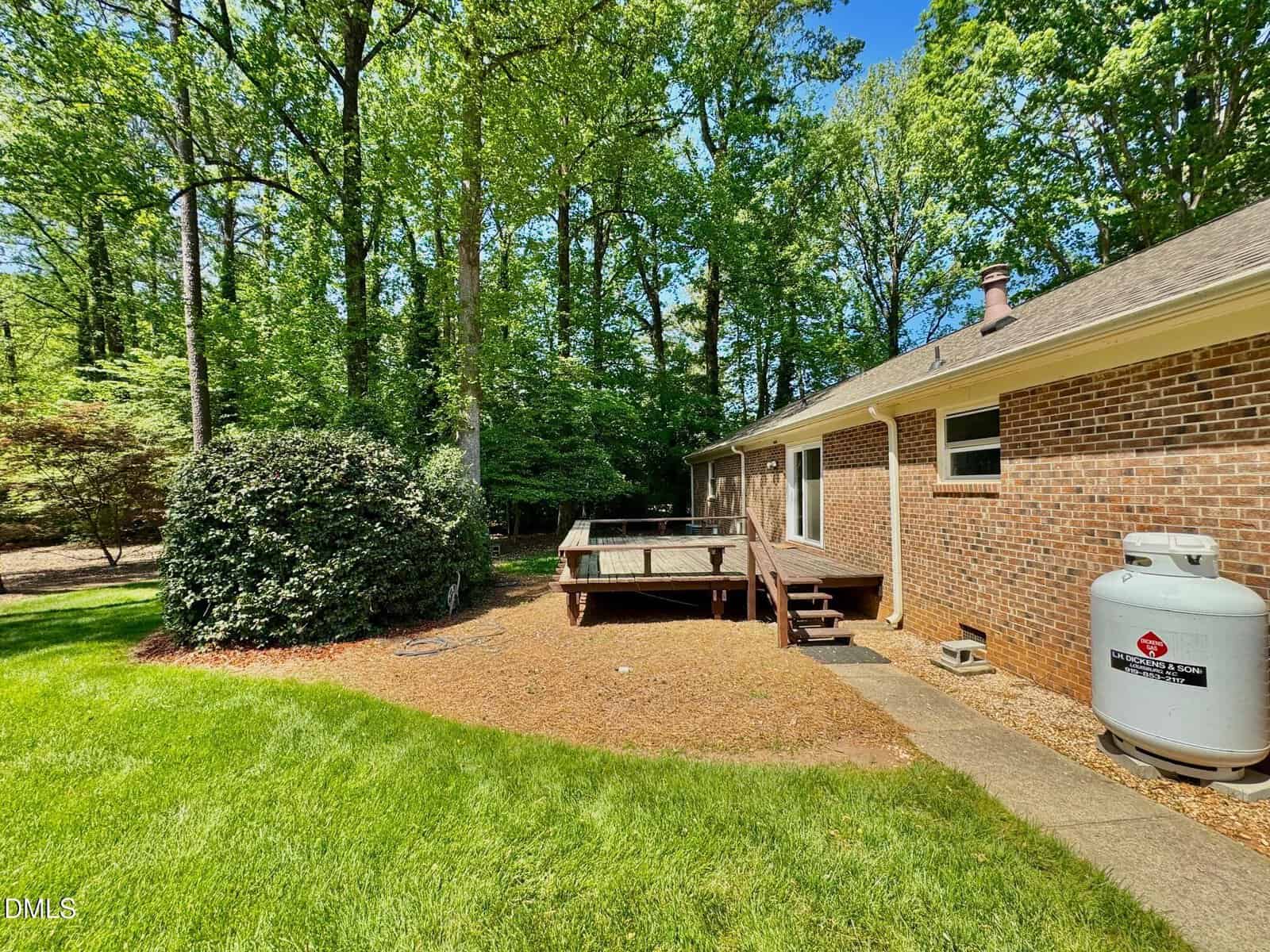 Backyard with green grass, large bushes, and tall trees. A wooden deck is attached to a brick house, with stairs leading down. A propane tank sits beside the house. The scene is bright and sunny.