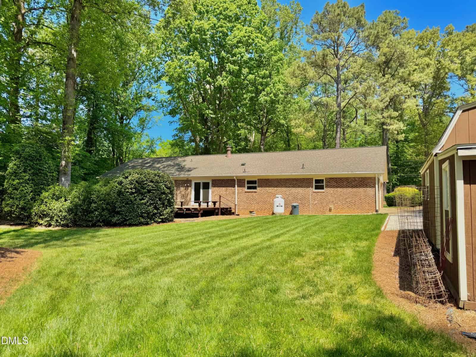 A brick single-story house with a sloped roof sits among tall trees. The backyard features a large, well-kept lawn, a small wooden deck, bushes, and a storage shed on the right side under a clear blue sky.