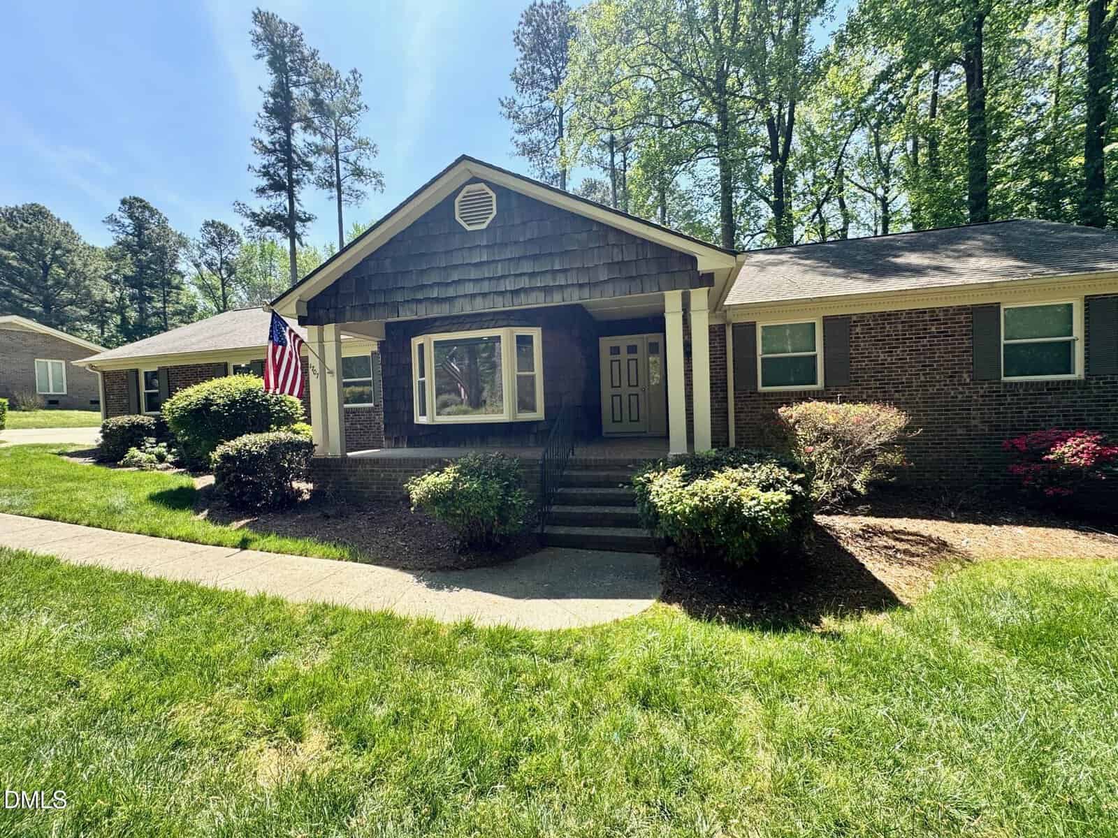 Charming single-story brick house at 1707 Peace Street in Henderson, featuring a covered front porch, double front doors, and a bay window. An American flag hangs by the entrance, with a landscaped yard and tall trees surrounding the home.