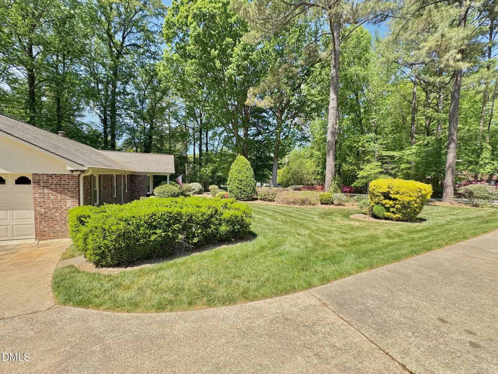 A well-maintained front yard with neatly trimmed bushes, green grass, tall trees, and a partial view of a brick house. A concrete driveway curves along the landscaped area under a clear sky.