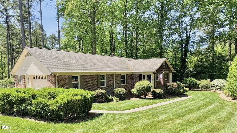 Single-story brick house with a gable roof, attached garage, and American flag on the front porch, surrounded by well-kept bushes and a green lawn, backed by tall trees and woods.