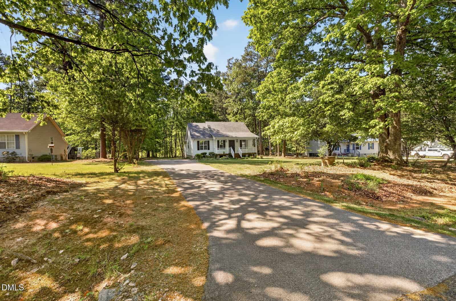 A small white house with a front porch sits at the end of a paved driveway, surrounded by large green trees and a grassy yard on a sunny day. Two neighboring houses are visible on either side.