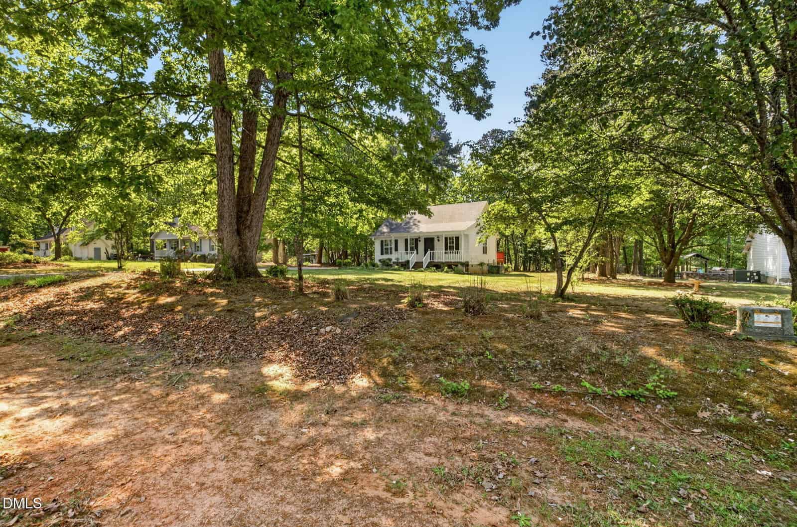 A white house with a front porch is surrounded by tall trees and greenery, set back from a dirt and leaf-covered yard on a sunny day. Another house and a small shed are visible in the background.