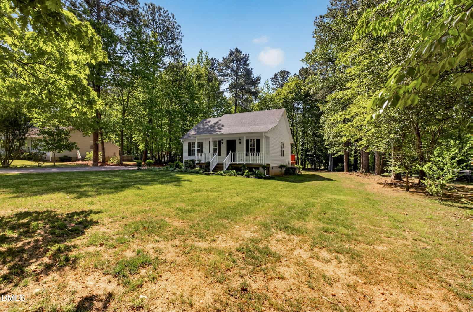 A small white house with a front porch is surrounded by green grass and tall trees under a bright blue sky.